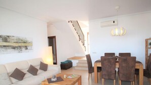 En moderne stue med sofa, sofabord og spisebord med stole. En trappe er synlig i baggrunden, der fører op til en anden etage.