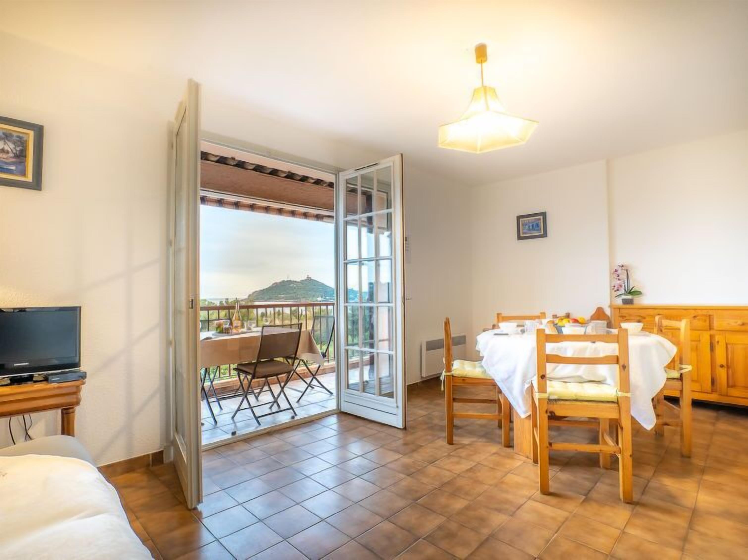 Et værelse med spiseplads, tv og en balkon med udsigt til en bakke.
