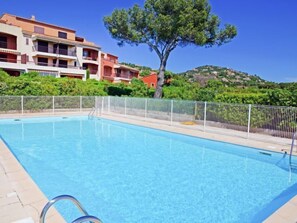 A swimming pool with a metal fence, surrounded by greenery and a building with balconies.