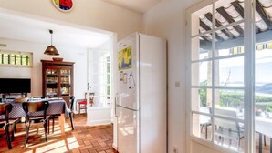 A kitchen with a refrigerator, a dining table with chairs, a clock, and a window with a view. - Maison La Galine by Interhome (Agay Saint Raphael)