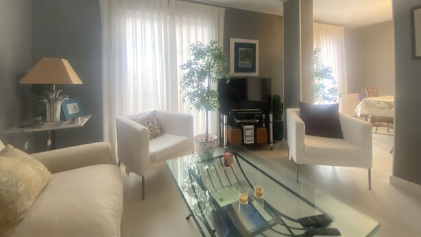 Uma sala de estar com uma mesa de centro de vidro, duas poltronas brancas, um sofá, uma televisão e uma planta.