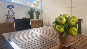 Uma mesa de madeira com um laptop, um vaso com maçãs verdes e cactos decorativos numa varanda com vista para edifícios.