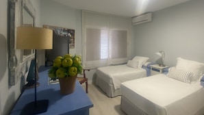 Quarto de hotel com duas camas, uma mesa com abajur, uma planta em vaso e uma janela com persianas.