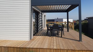Una terraza de madera con una pérgola, una mesa con sillas y vistas a edificios cercanos.