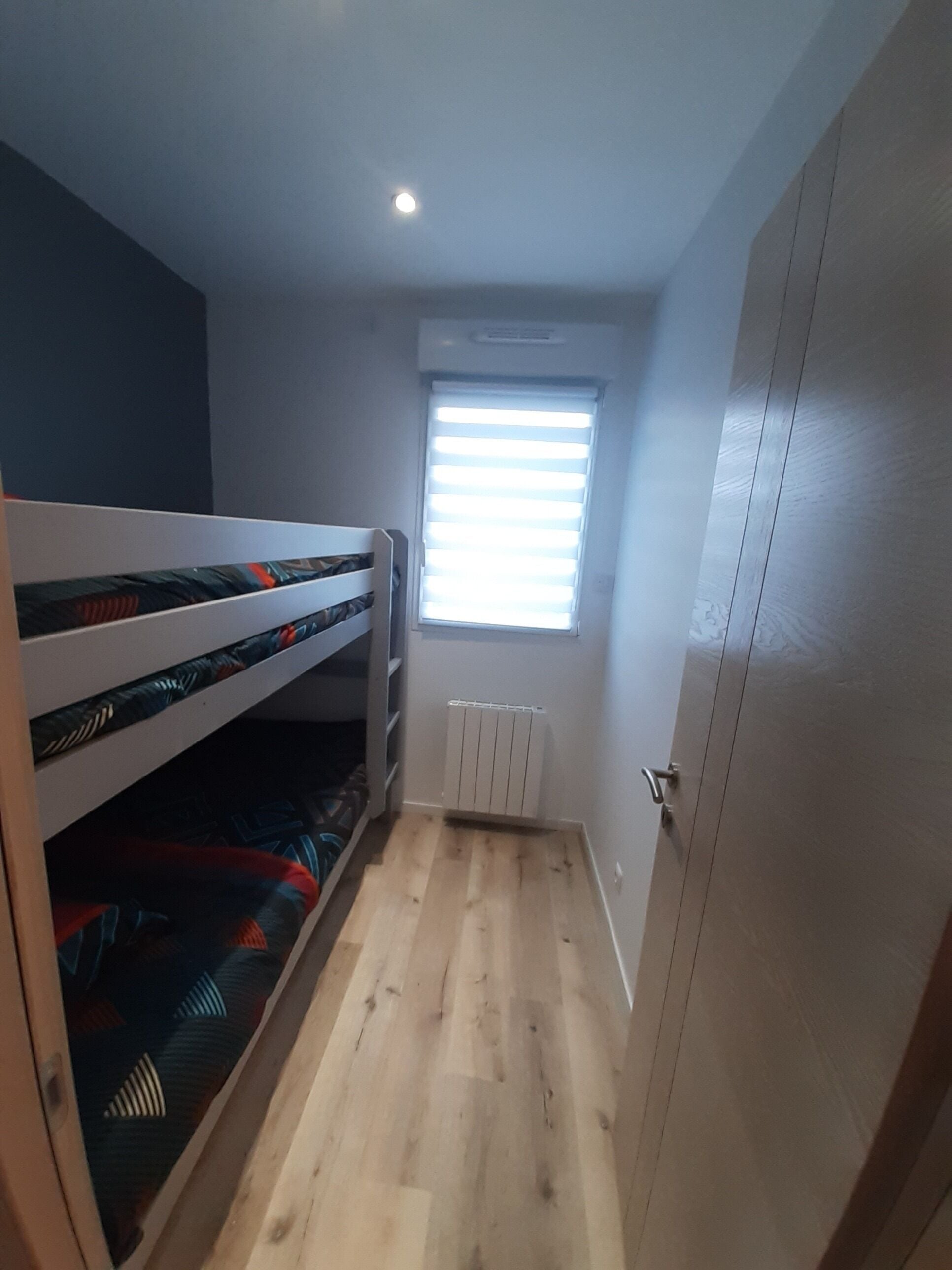 A small room with a bunk bed, a window with blinds, a radiator, and a door.