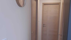 Un pasillo estrecho con suelo de madera, un reloj en la pared y una puerta cerrada al fondo.