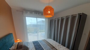 Un dormitorio con una cama grande, un cabecero, una lámpara colgante anaranjada y vistas a través de una ventana con cortinas.