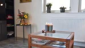 Una sala de estar moderna con una mesa de centro de vidrio, una mesita auxiliar de madera con velas y un florero con flores.