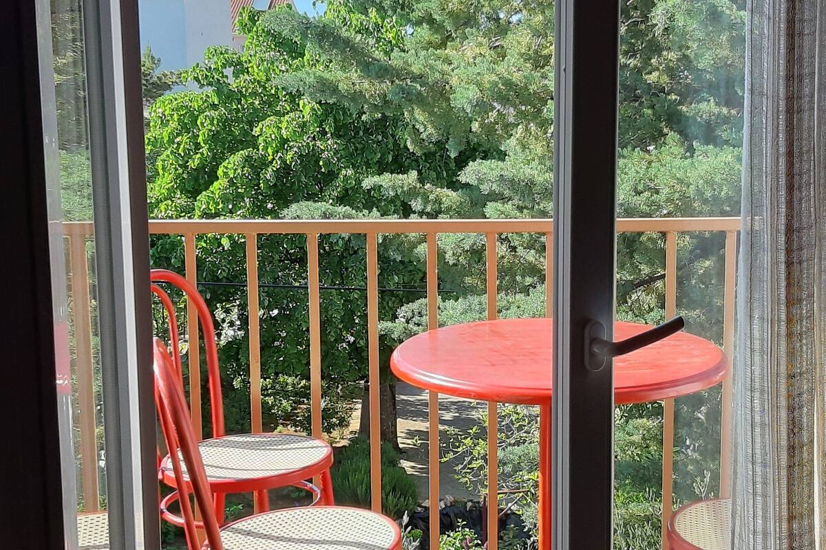 A balcony with a red table and two red chairs, surrounded by greenery.