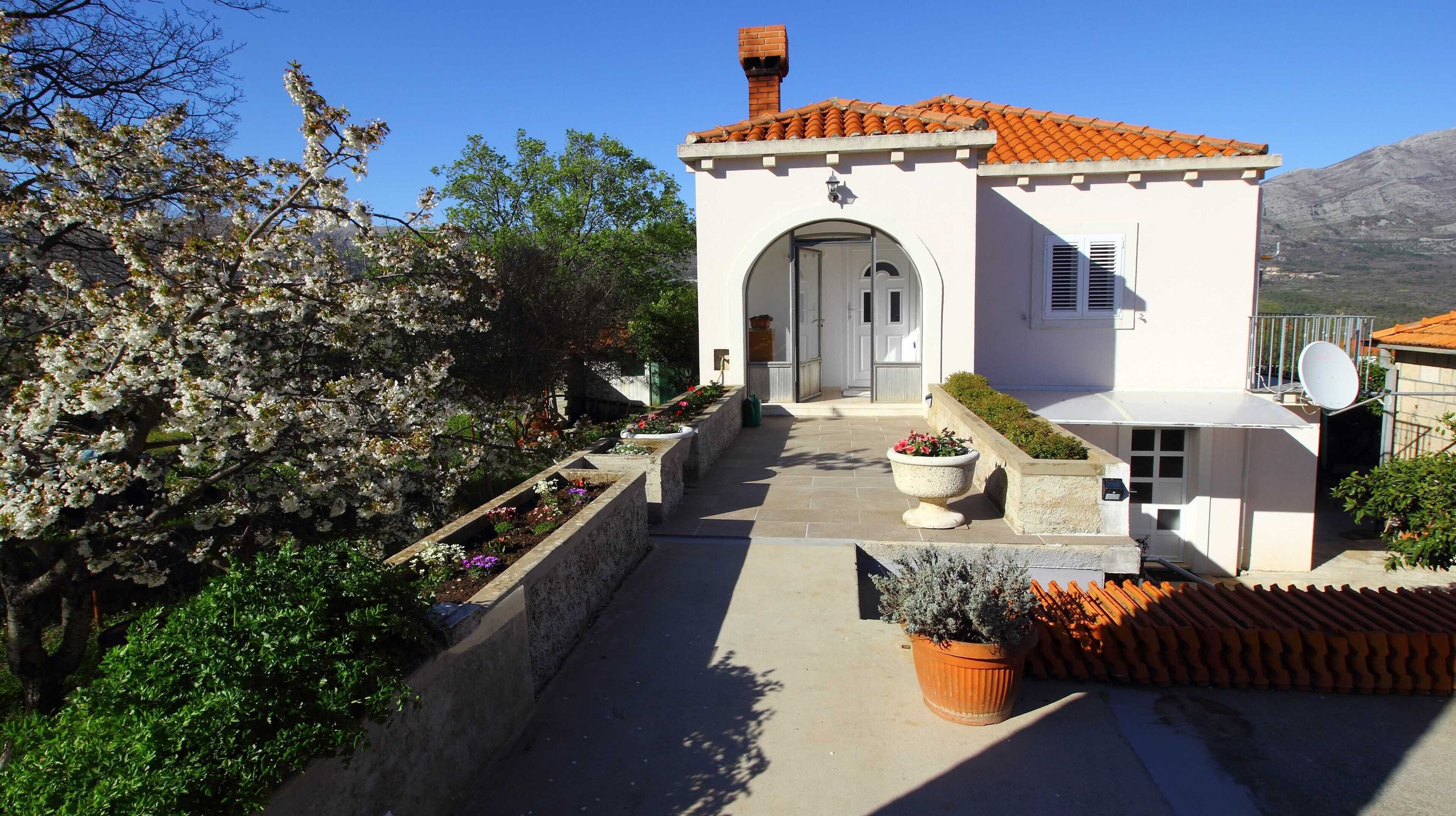 A small building with a tiled roof, a central entrance, and a garden with flowering plants.