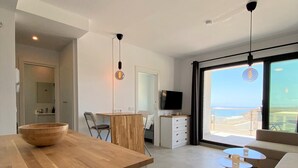 Une cuisine moderne avec un îlot central en bois, un coin repas avec une table et des chaises, un téléviseur et une grande fenêtre donnant sur la mer.