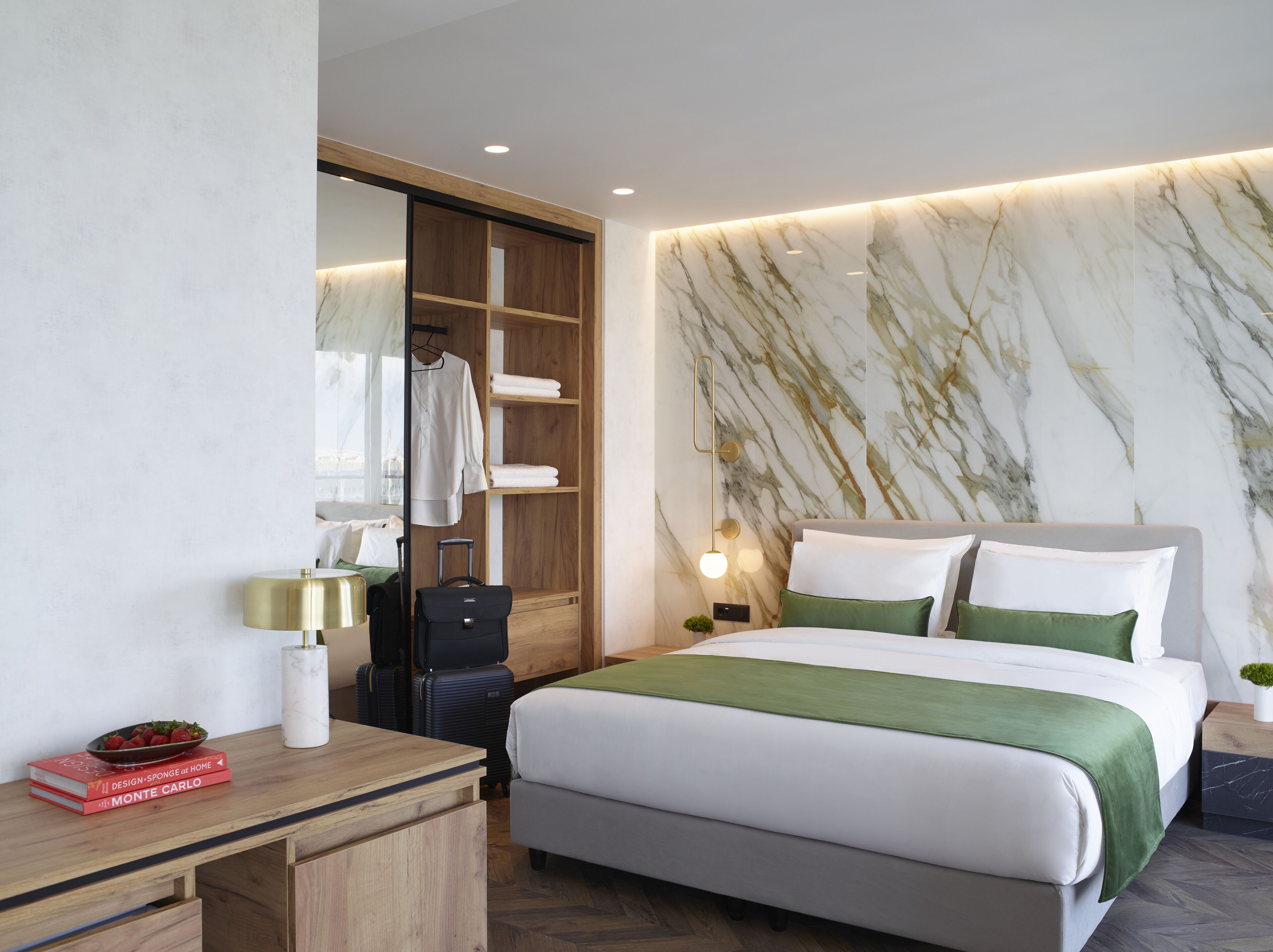 Una habitación de hotel moderna con una cama grande, un armario de madera, un escritorio con lámpara y una pared con estampado de mármol.