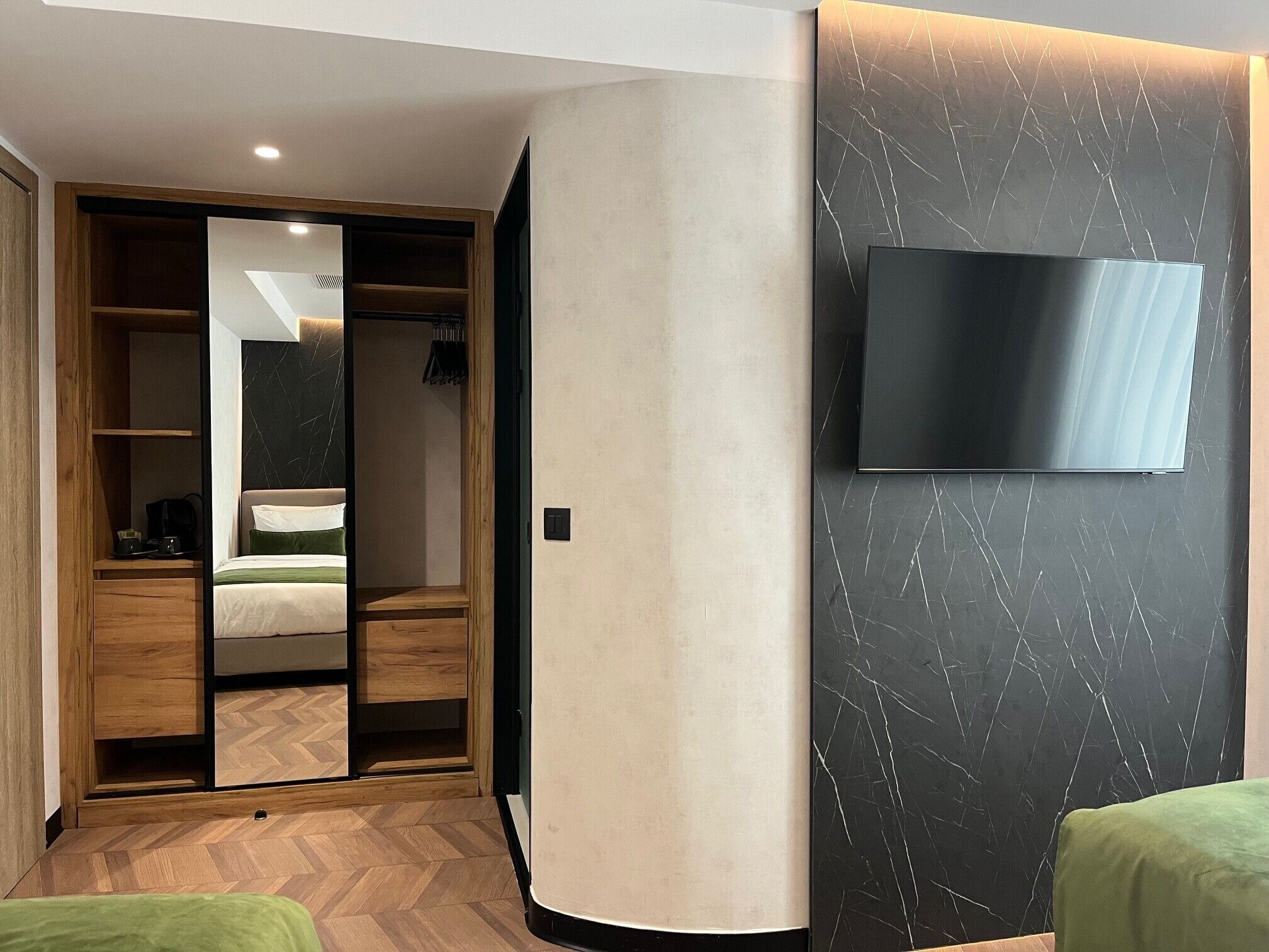A hotel room with a wooden wardrobe, a bed with green bedding, a television mounted on a marble-patterned wall, and a herringbone-patterned floor.