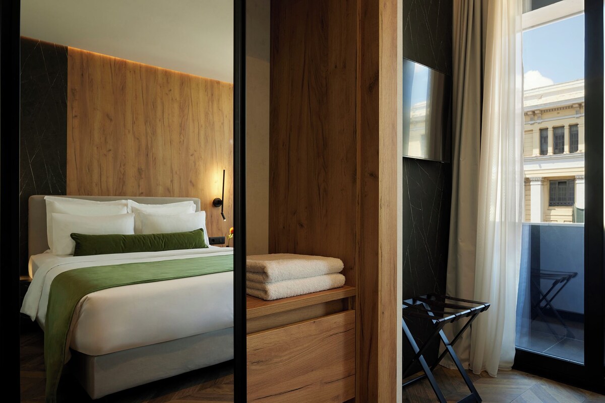 A modern hotel room with a wooden accent wall, a bed with green bedding, a small bedside table with a lamp, and a view of a building through a large window.