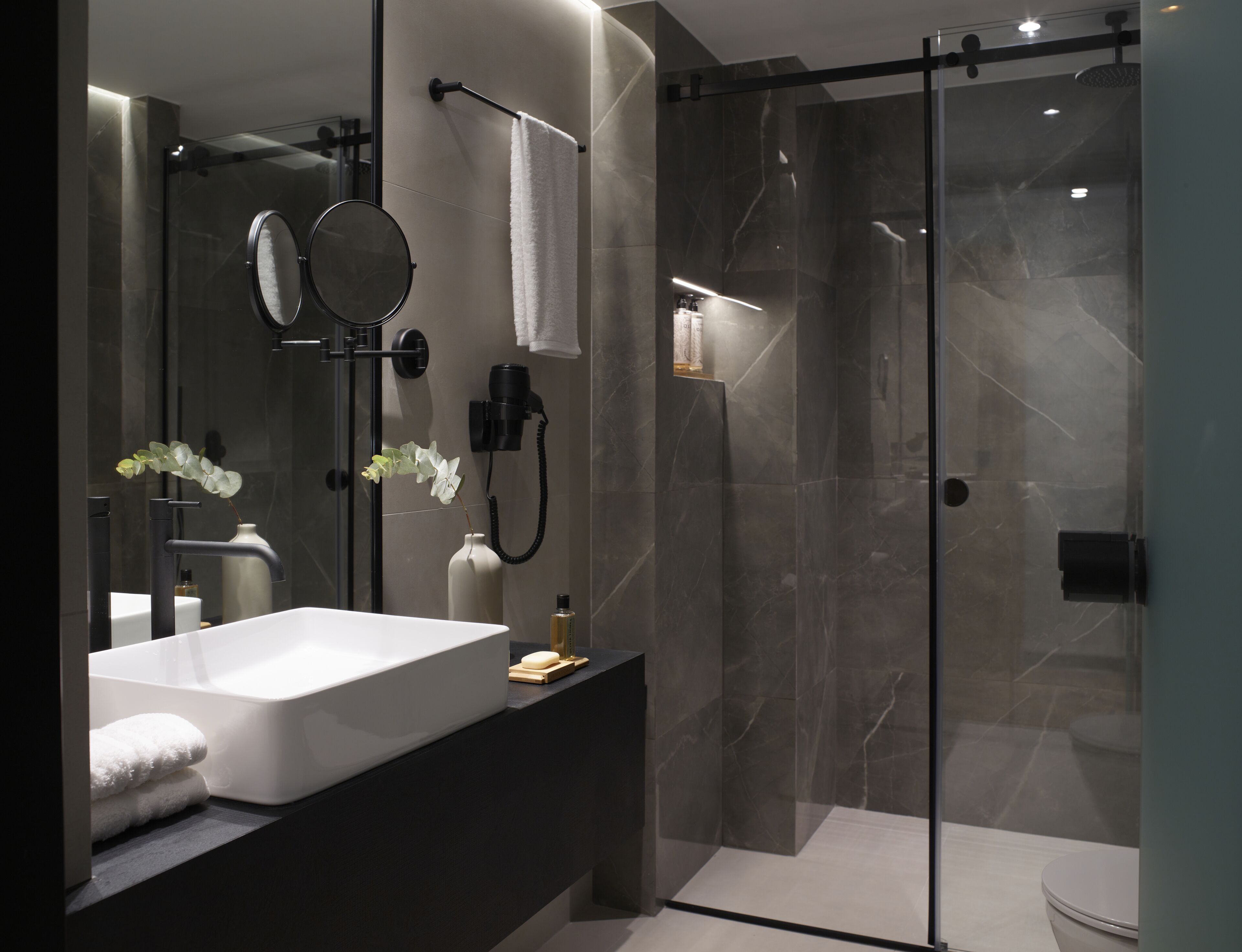 A modern bathroom with a large white bathtub, a glass-enclosed shower, and a vanity with a mirror and a vase of flowers.