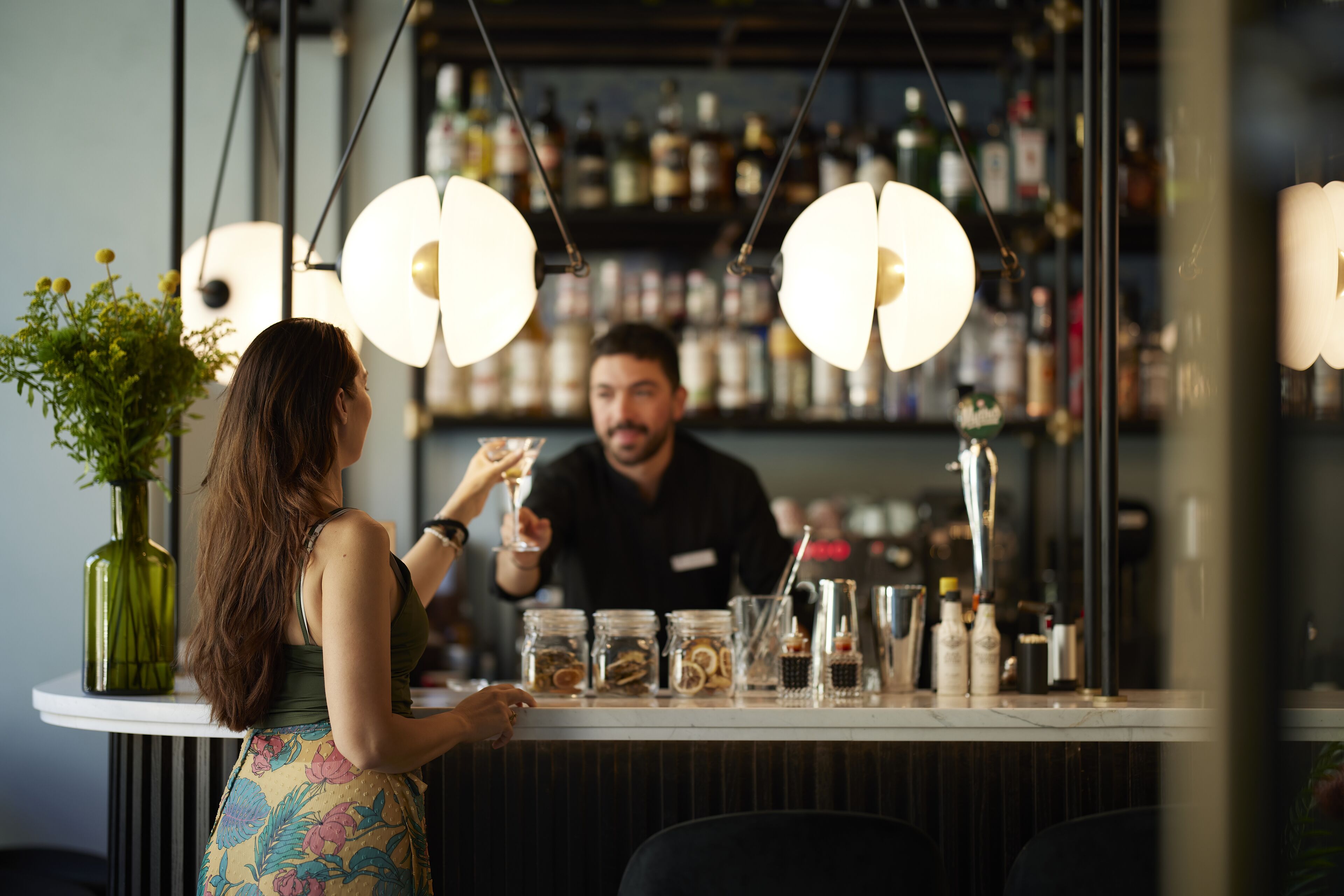 A bar with a bartender, a counter with various bottles and jars, and hanging pendant lights.