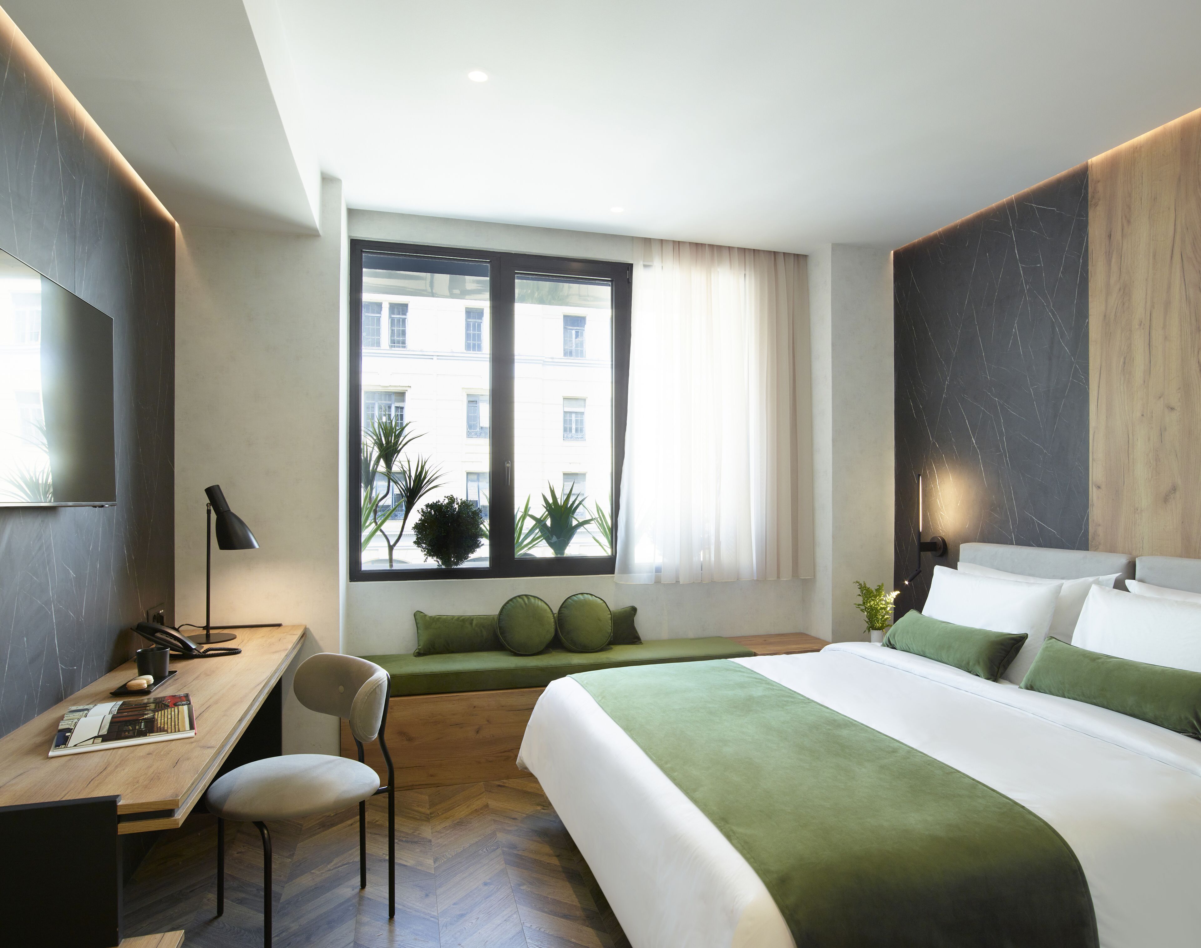 A modern hotel room with a large bed, a desk, a chair, and a window with a view of buildings.