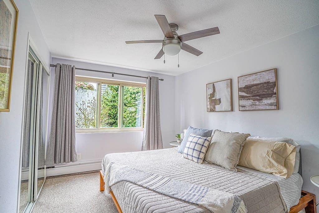 Un dormitorio con una cama, un ventilador de techo, una ventana con cortinas y dos cuadros en la pared.