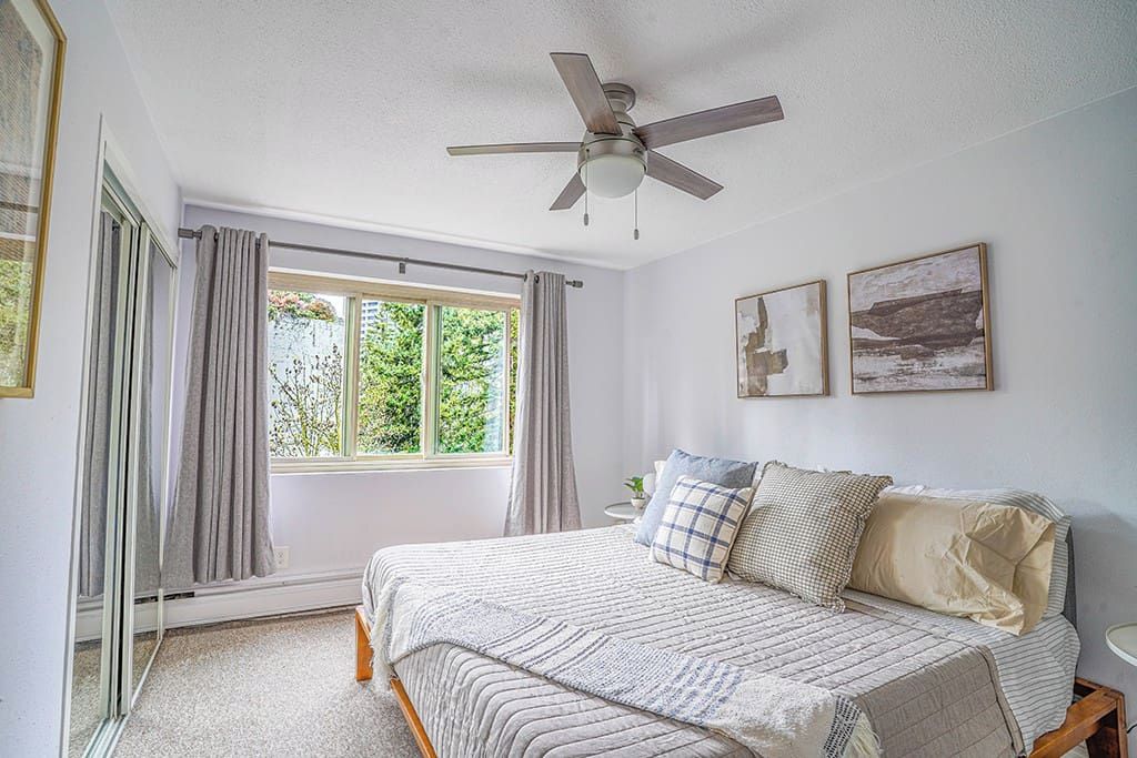 Une chambre à coucher avec un lit, un ventilateur de plafond, une fenêtre avec des rideaux et deux tableaux encadrés accrochés au mur.