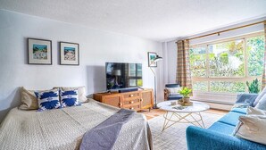 Un salón bien iluminado con una cama grande, un sofá azul, un televisor y una ventana con cortinas.