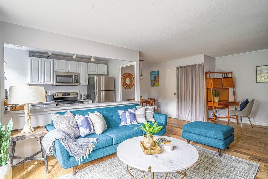 A modern living room with a blue sofa, a round coffee table, and a kitchen area with white cabinets and appliances.