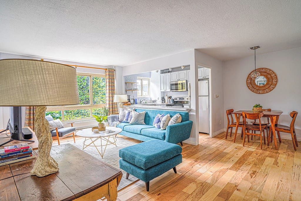 A modern living room with a blue sofa, a coffee table, and a lamp. There is a kitchen in the background with white cabinets and a dining area with wooden chairs.