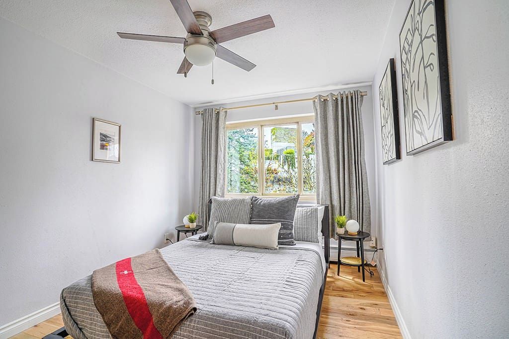 Une chambre à coucher avec un lit, un ventilateur de plafond, une fenêtre avec des rideaux, une petite table avec une plante et des œuvres d’art encadrées accrochées au mur.