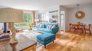 Una sala de estar moderna con un sofá azul, una mesa de centro y una lámpara. Se observa una cocina al fondo con armarios blancos y un comedor con sillas de madera.