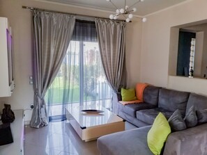 Ein modernes Wohnzimmer mit einem grauen Sofa, einem Glastisch und Blick durch große Fenster mit grauen Vorhängen.