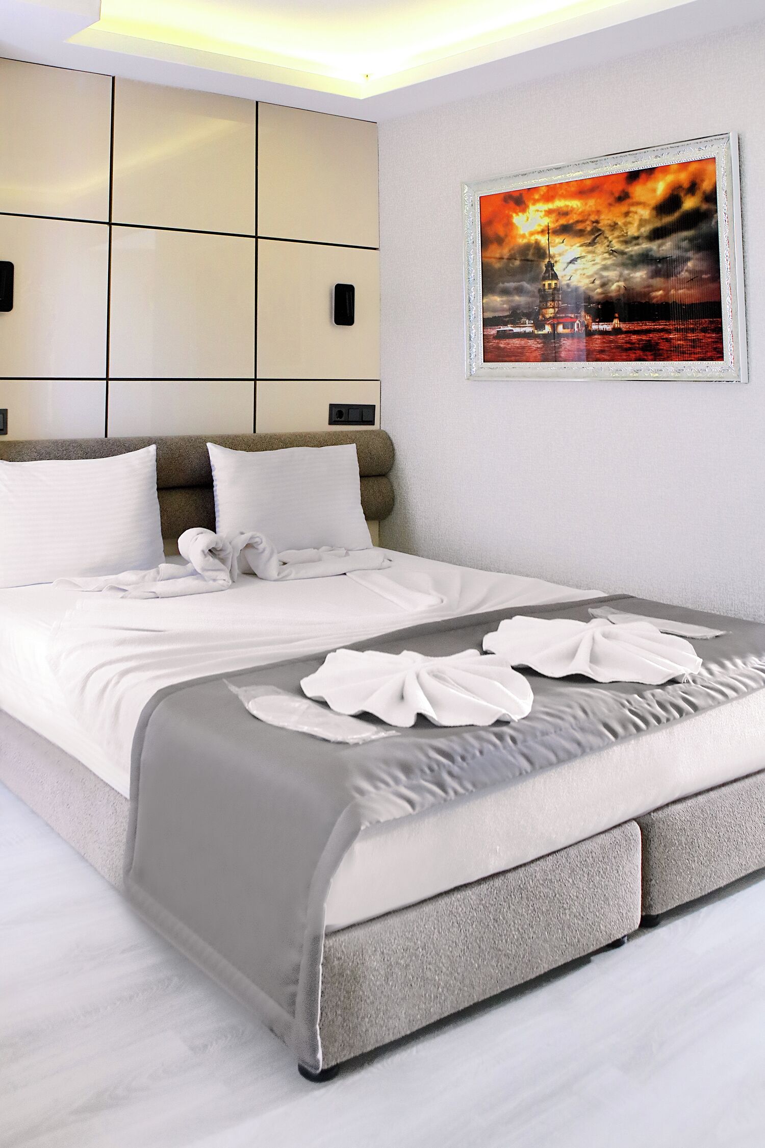Una habitación de hotel con una cama grande, un cabecero con compartimentos de almacenamiento, un cuadro enmarcado de un atardecer y una cama bien hecha con toallas.