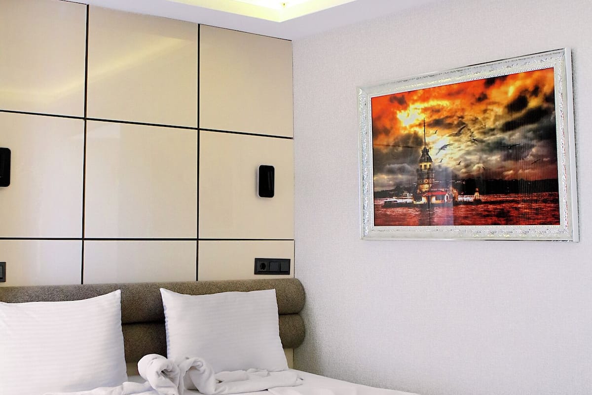 Una habitación de hotel con una cama grande, un cabecero con compartimentos de almacenamiento, un cuadro enmarcado de un atardecer y una cama bien hecha con toallas.