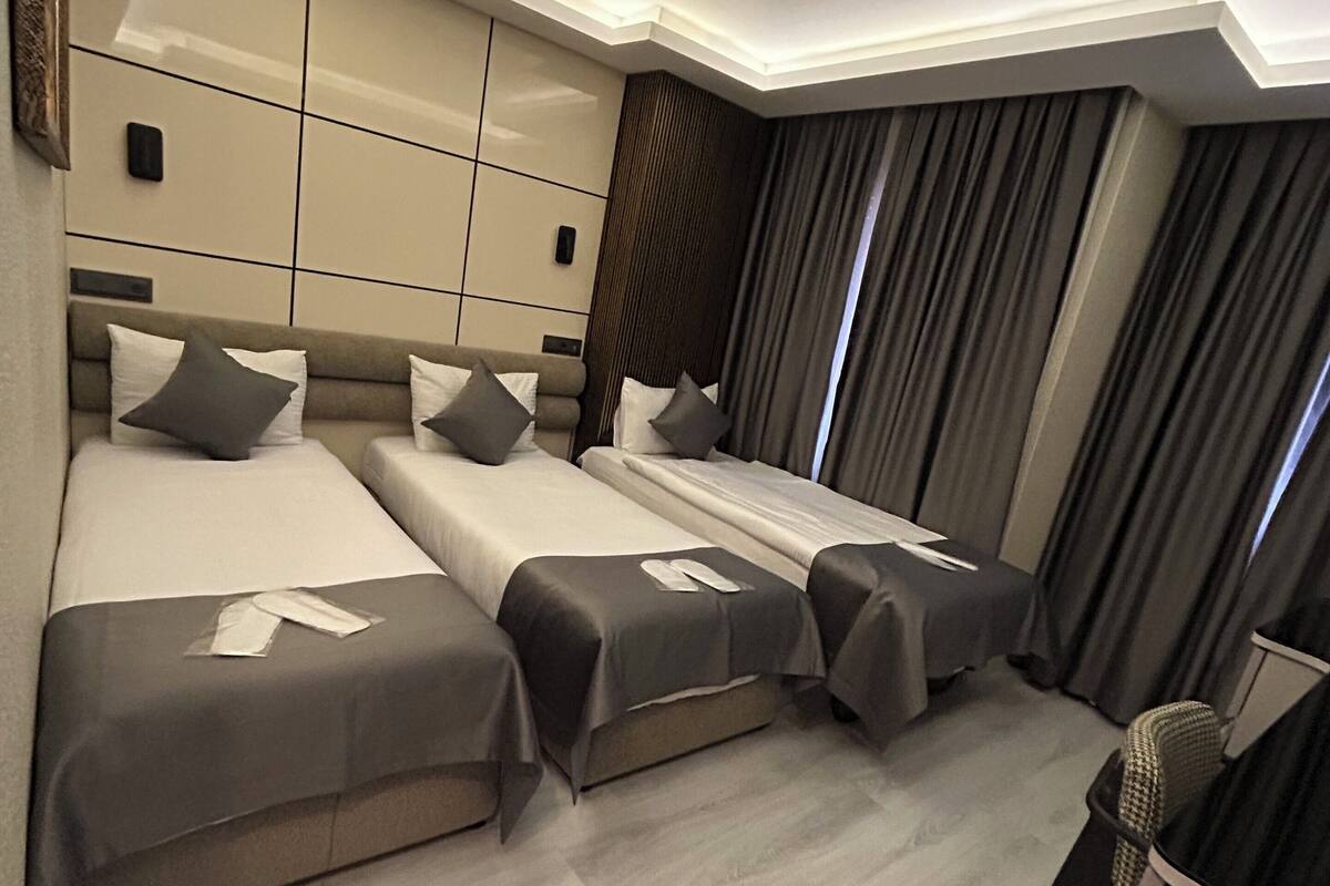 A hotel room with a modern design, featuring a large bed, a smaller bed, and a sofa bed. The room has a neutral color scheme with beige and gray tones.