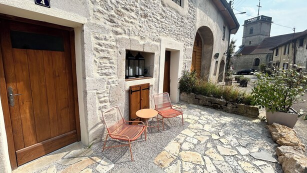 A stone building with a wooden door, a small patio with a table and chairs, and a view of a church in the background.
