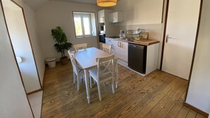 A compact kitchen with white cabinets, a wooden dining table, and chairs. There is a potted plant and a hanging light fixture.