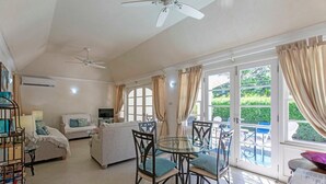 A spacious living room with a glass dining table, comfortable seating, a sofa, and a television. Large windows offer a view of the outdoors. - The Falls Villa No. 1 | Elegant Caribbean Retreat in Holetown (Holetown)