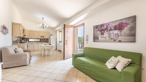 En rummelig opholdsstue med en grøn sofa, en beige sofa og et spisebord. Et køkken ses i baggrunden med træskabe og en lysekrone.