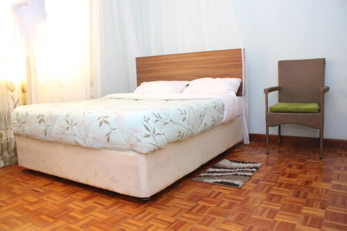 Un dormitorio con cama de cabecera de madera, una butaca verde acolchada y una alfombra estampada sobre un suelo de parqué.