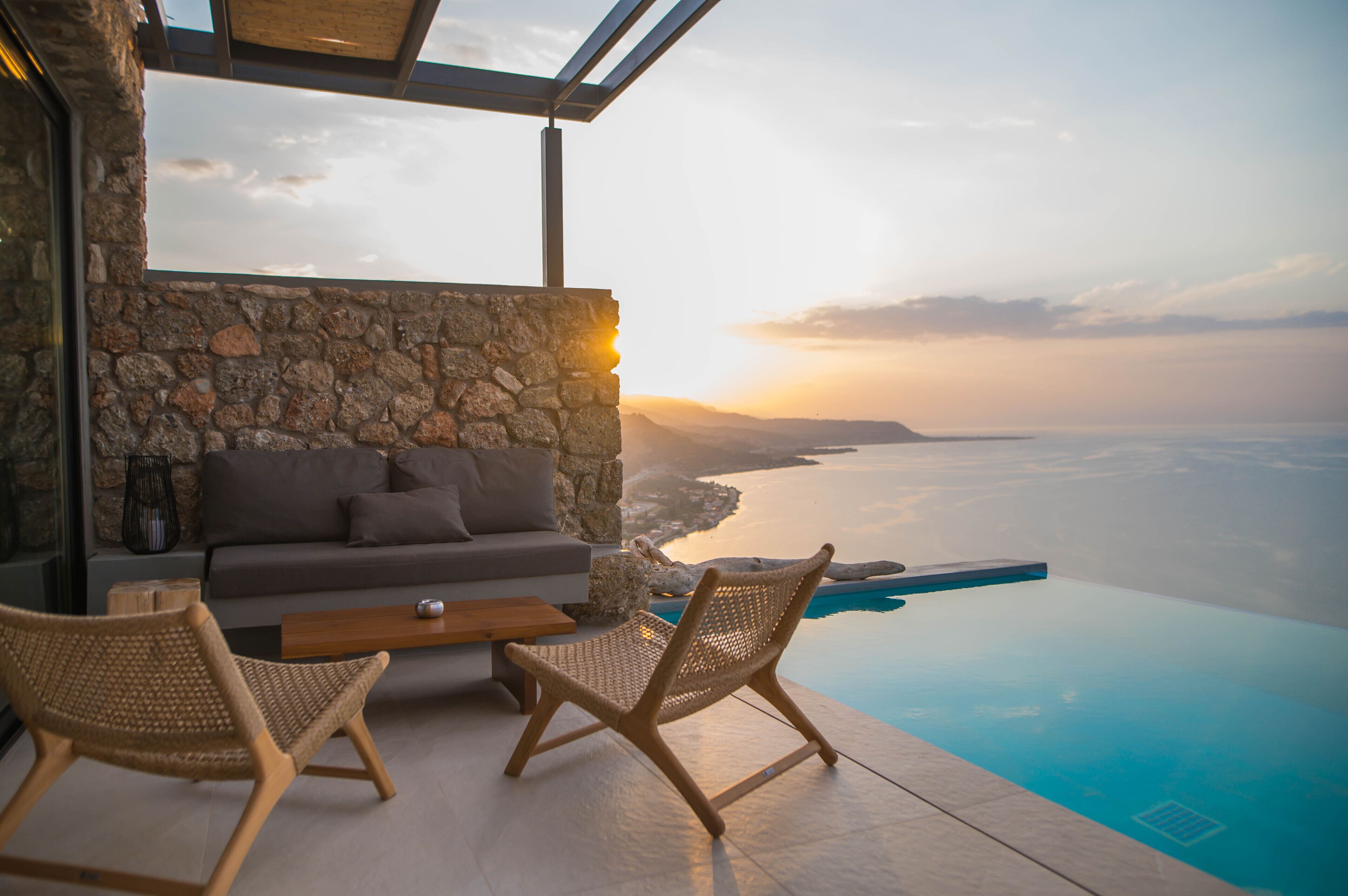 A modern outdoor seating area with a stone wall, a sofa, and two wicker chairs overlooking a clear blue pool and a scenic view of the sea at sunset.