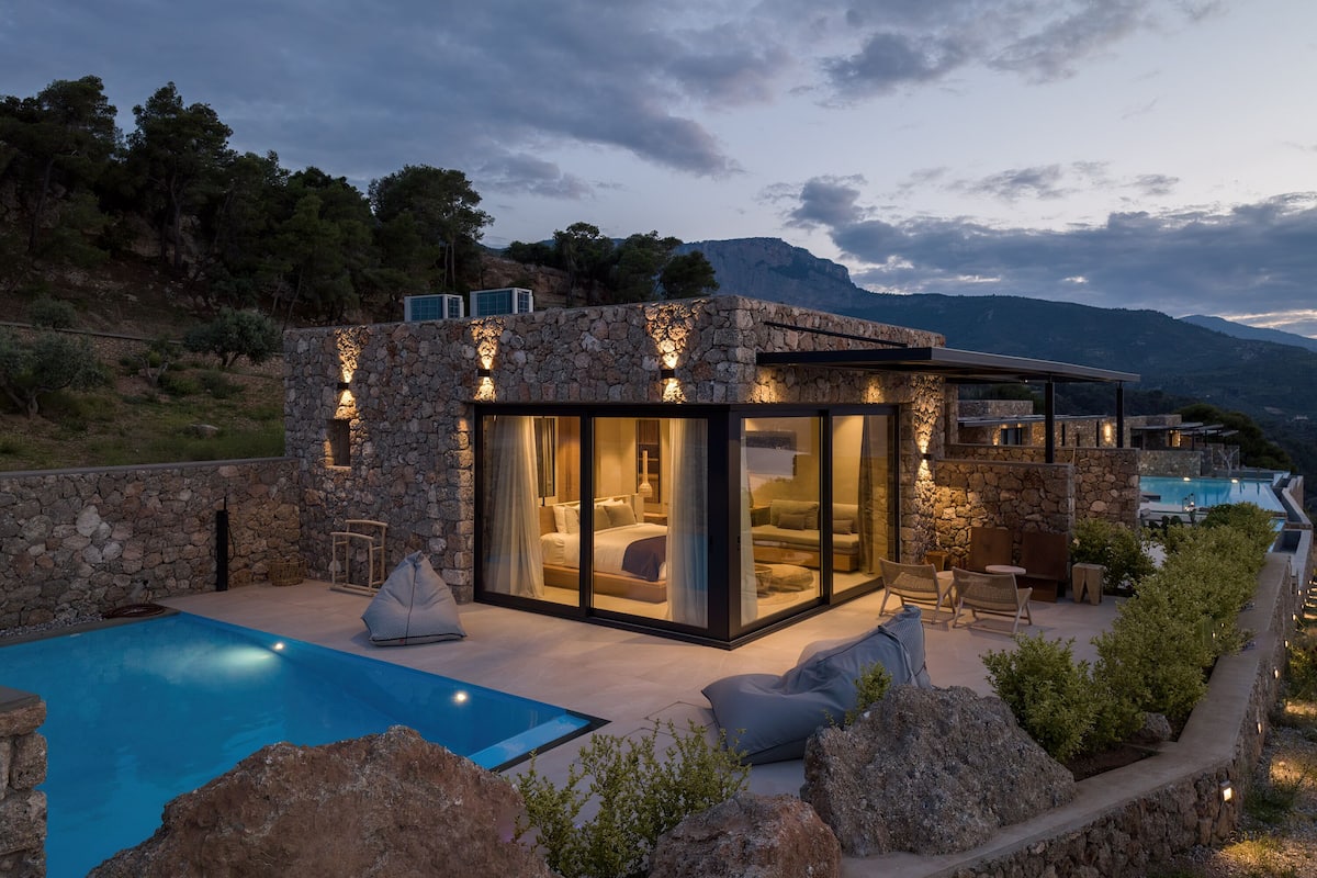 Una villa moderna con fachada de piedra, un dormitorio con paredes de cristal y una piscina, con vistas al paisaje circundante.