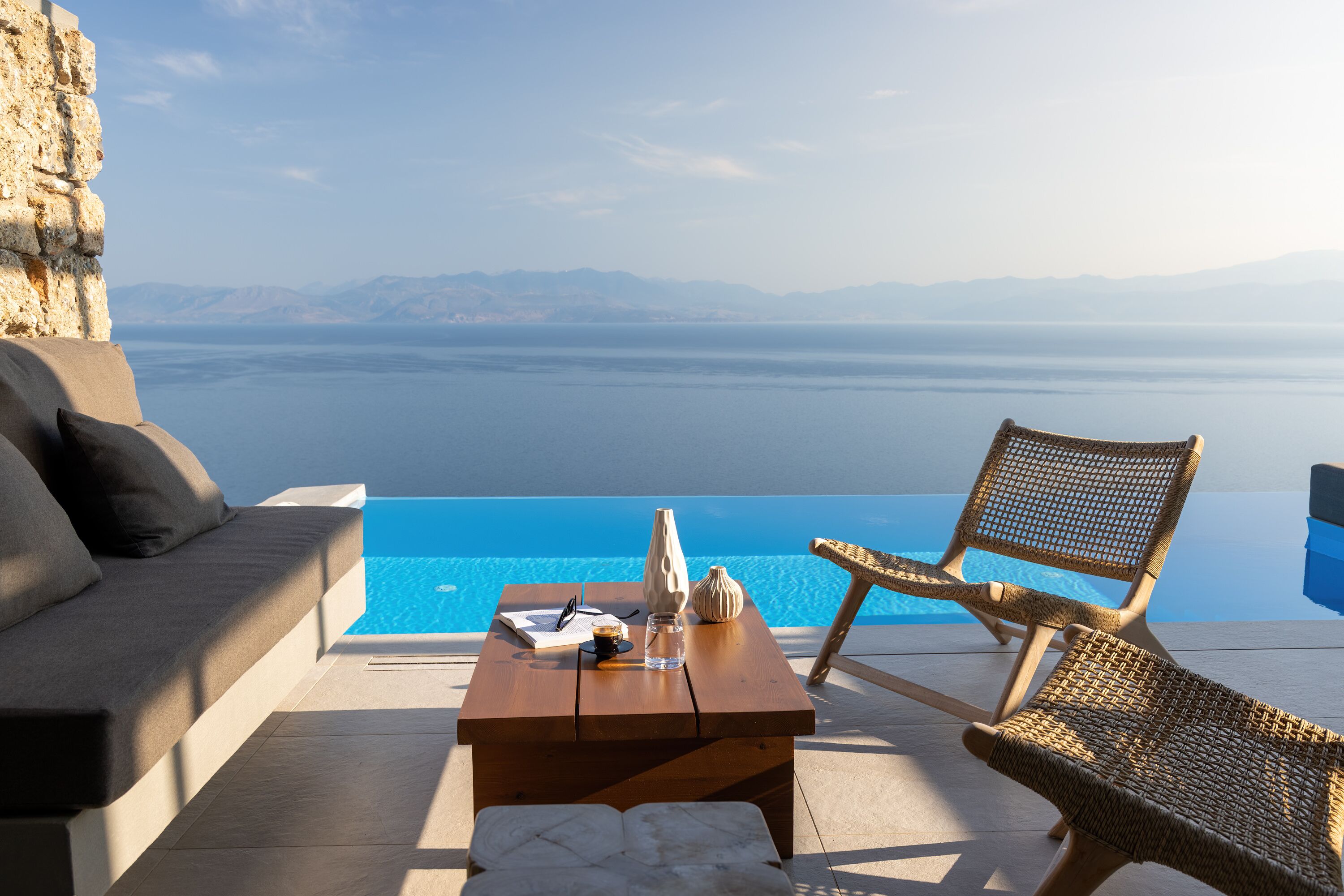 A poolside area with a wooden table, wicker chairs, and a sofa overlooking a clear blue sea with distant mountains.