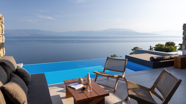 Ein Pool ohne Rand mit Blick auf Meer und Berge, ein Holztisch mit einer Vase und Korbstühle.