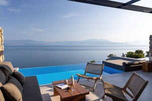 An infinity pool with a view of the sea and mountains, a wooden table with a vase, and wicker chairs.