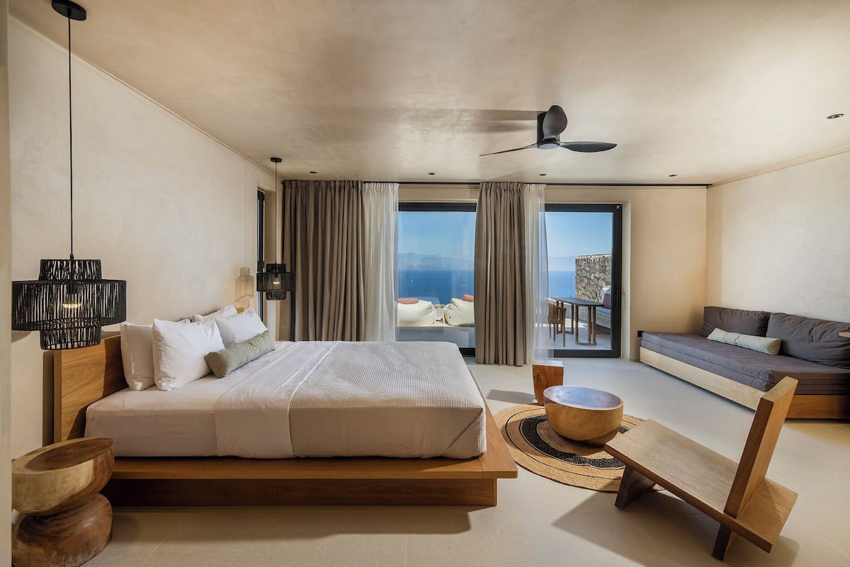 Un dormitorio moderno con una cama grande, una mesita de noche de madera, un banco de madera y vistas al mar a través de amplias ventanas.