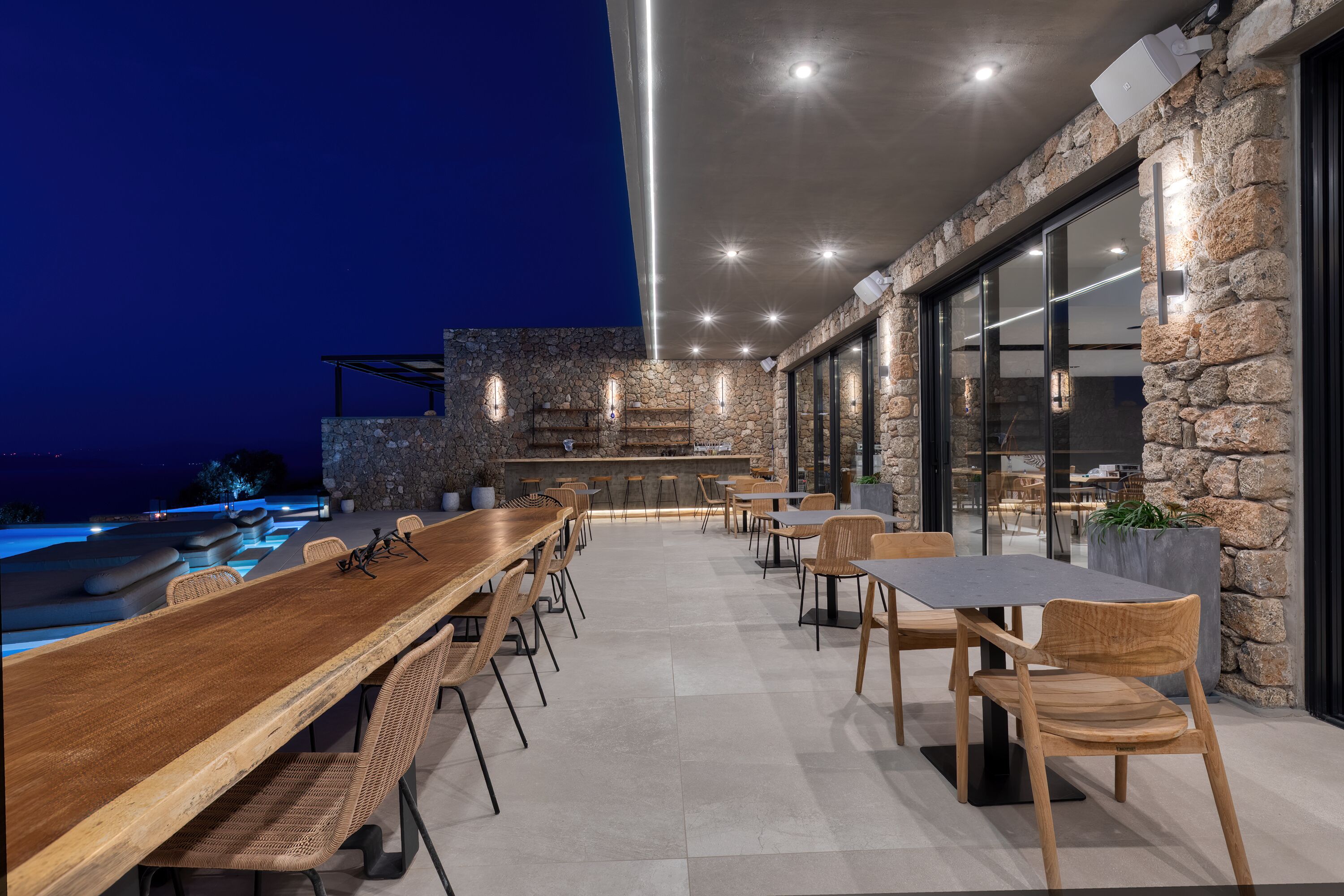 An outdoor dining area with a long wooden table and chairs, a stone wall, and a view of the pool.