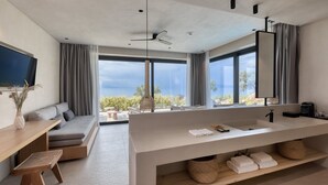 Ein modernes Hotelzimmer mit großem Fenster, einer Sitzecke, einem Holztisch, einem großen Waschbecken und einem Deckenventilator.