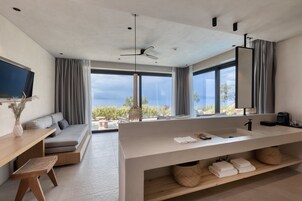 A modern hotel room with a large window, a sofa, a wooden table, a large sink, and a ceiling fan.