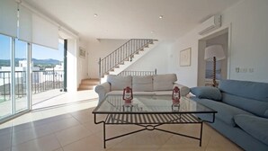 A modern living room with a glass coffee table, a blue sofa, a staircase, and a balcony with a view of the city. - Casa Can Alou, authentic Mallorquin house nestled in the village of Porto Colom (Felanitx)