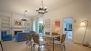 A dining area with a glass table and wicker chairs, a chandelier, and a blue sofa in a well-lit room with white walls and a fireplace. - Casa Can Alou, authentic Mallorquin house nestled in the village of Porto Colom (Felanitx)
