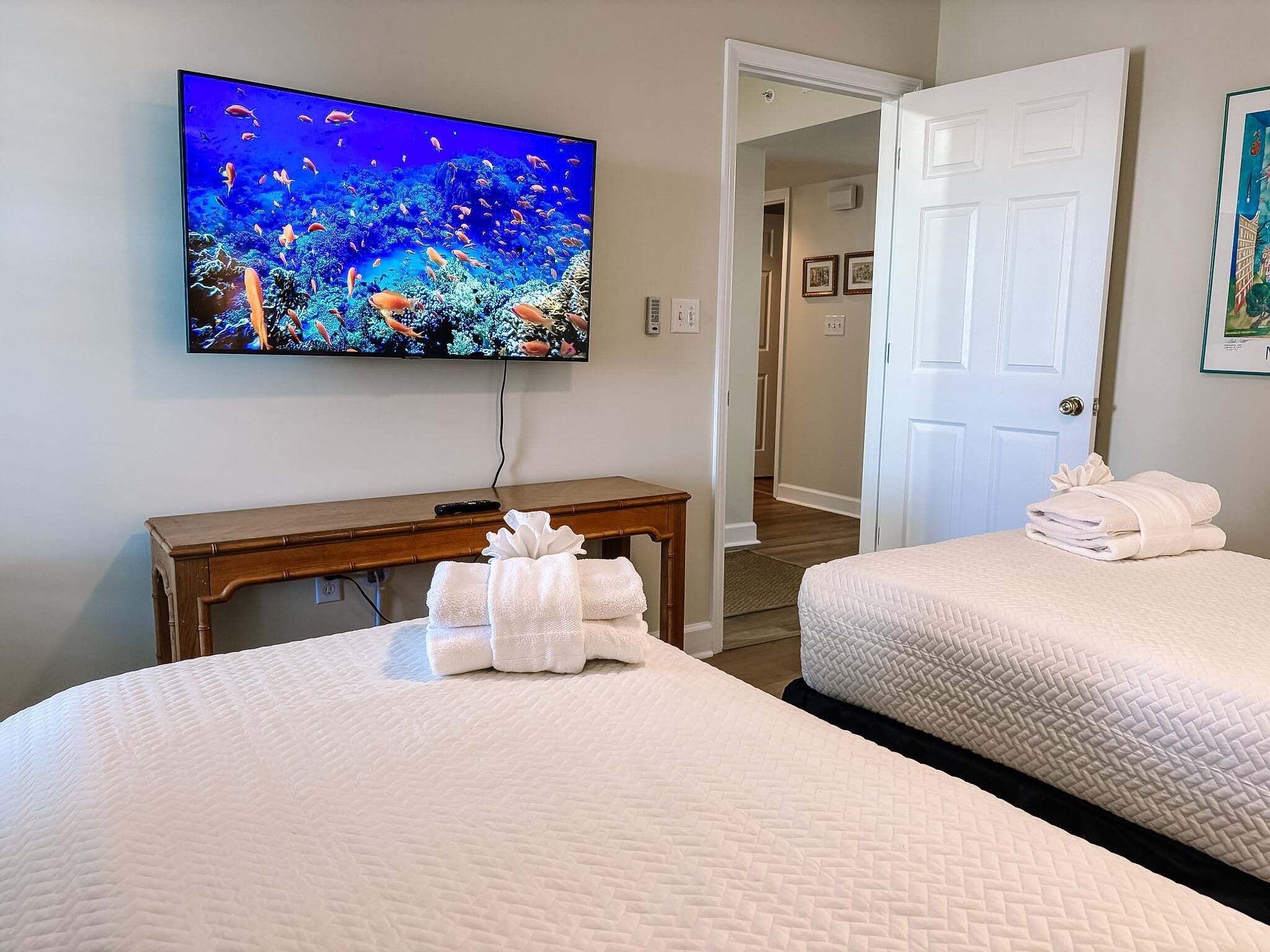 Een hotelkamer met twee bedden, een televisie waarop een onderwaterscène wordt weergegeven, een houten tafel met handdoeken, en een deur die naar een andere kamer leidt.