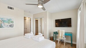 Ein Hotelzimmer mit einem Bett, einem Schreibtisch, einem Stuhl, einem Deckenventilator, einem Fernseher und einem Bild an der Wand.